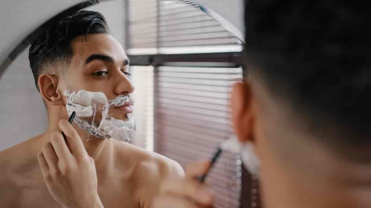 Mann med barberskum i fjeset som barberer seg med høvel mens han ser seg i speilet.