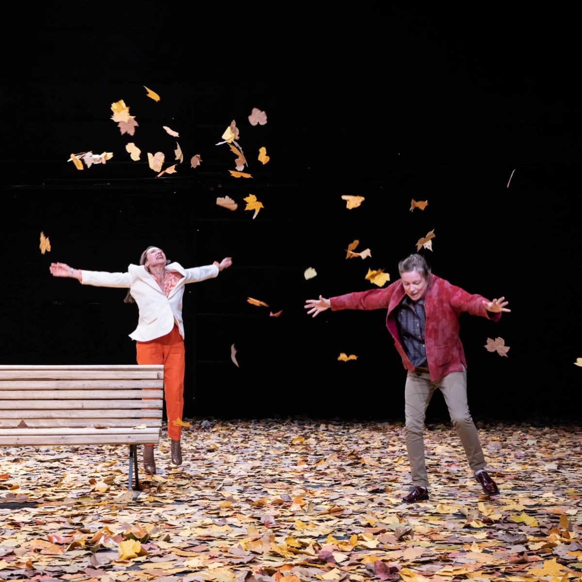 To smilende mennesker på en scene hopper og kaster høstløv opp i luften