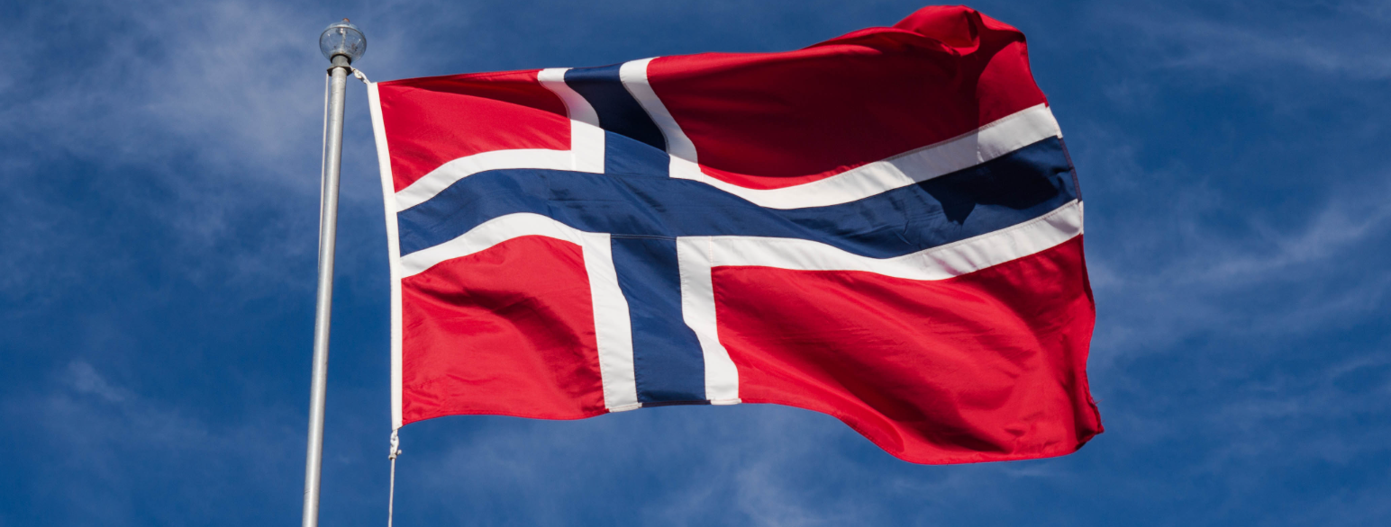 Et fotografi av et norsk flagg som vaier i vinden fra en hvit flaggstang. Himmelen er blå. 
