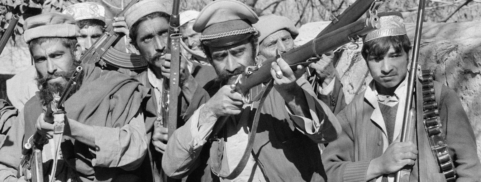 Fem-seks menn kledd i tradisjonelle afghanske klær poserer med våpen, en av dem har en offiserslue, antagelig sovjetisk.