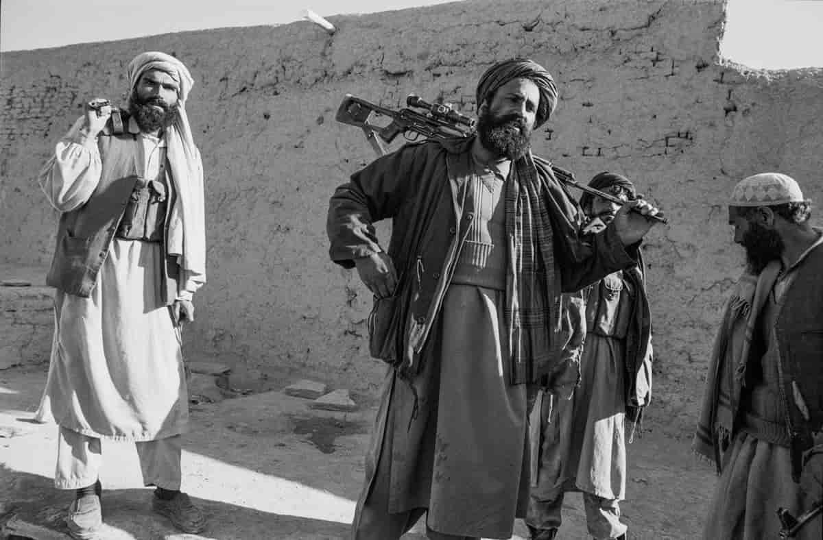 Tre menn i tradisjonelle afghanske klær, lange skjorter og vide bukser og turban, bærer våpen. 