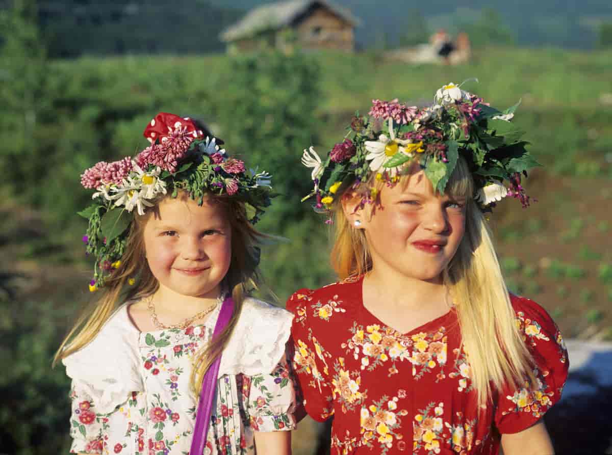 To jenter på cirka 7 og cirka 9 år med blomstrete kjoler og blomsterkranser i håret står utendørs i grønt gress. Det er sommertid og sol. 