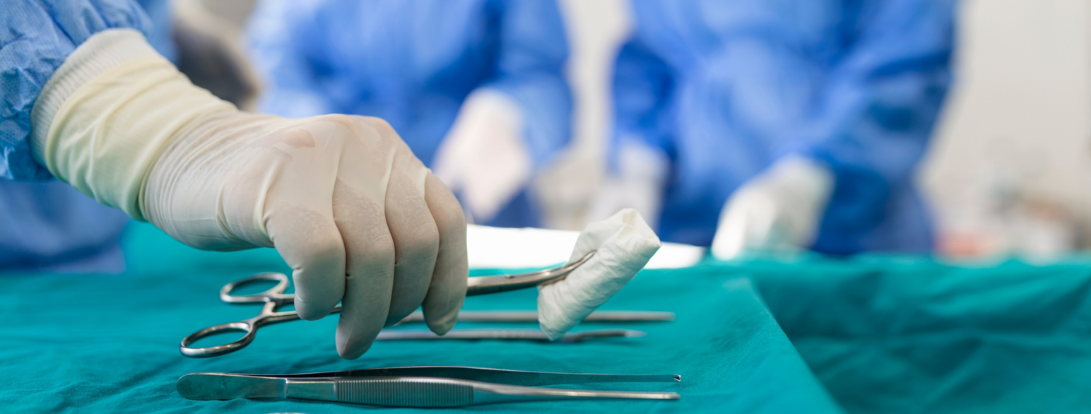 Bildet er fra en operasjonssal. En hånd med hansker plukker opp en metallklype fra en grønn duk. Klypen holder en kompress, en slags bomullspute som stopper blødninger. Bakover i bildet ser man to kirurger ved et operasjonsbord.