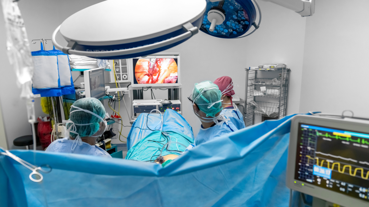 Helsepersonell står rundt ein pasient på eit operasjonsbord. To kirurgar held utstyr for kikholskirurgi. Små kamera inni kroppen til pasienten sender eit direktesendt opptak til ein skjerm ved fotenden av bordet, så kirurgane kan sjå kvar i kroppen dei skal skjera.