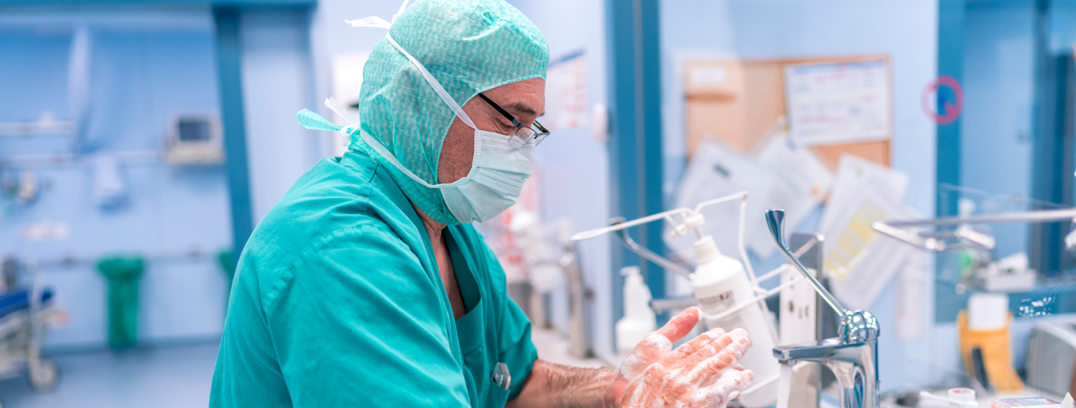 Ein mannleg kirurg står ved ein stor oppvaskkum utanfor operasjonssalen. Har har på seg grøne klede, ein heitte og munnbind. Han førebur seg til operasjon ved å vaska hendene grundig.