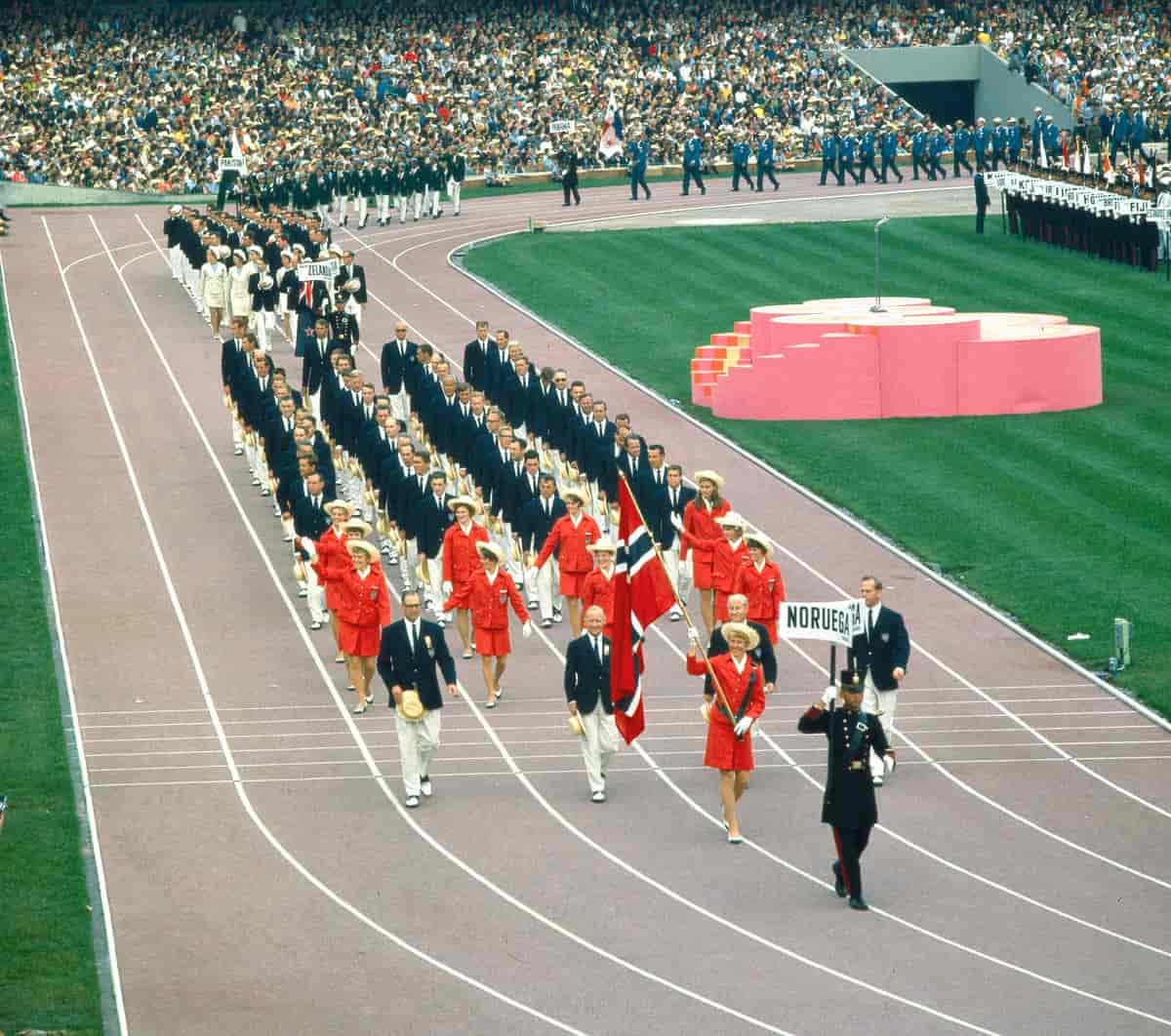 En gruppe idrettsutøvere marsjerer i tropp på en friidrettssarena, anført av en flaggbærer.