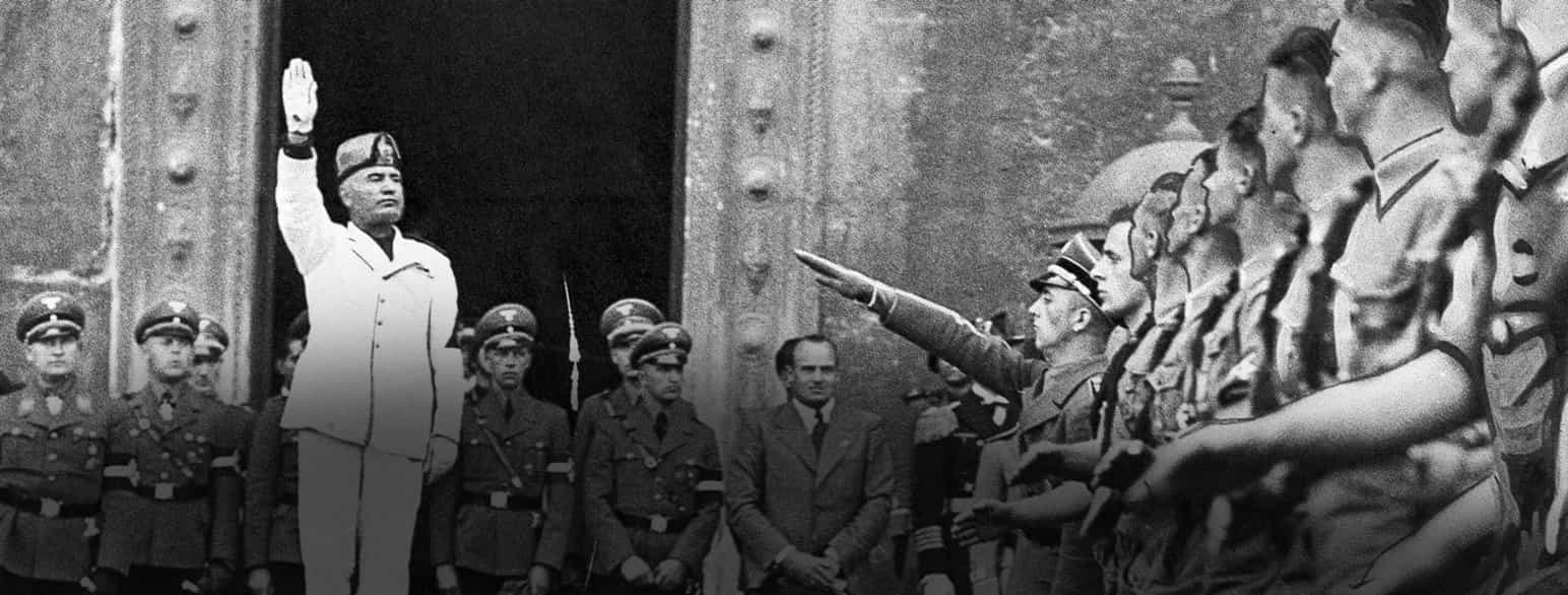 Svart-hvittfoto. Mussolini er kledd i hvit bukse og jakke og løfter armen. Bak ham står uniformskledde menn. Foran og til høyre i bildet er det marsjerende unge menn i uniformer med korte bukser og hakekors på ermet. 
