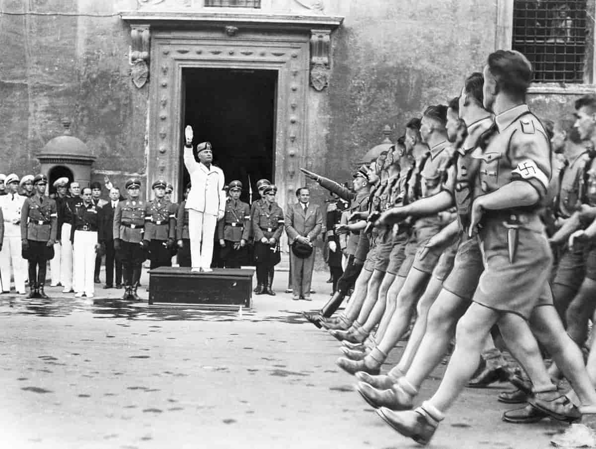 Svart-hvittfoto. Mussolini er kledd i hvit bukse og jakke og løfter armen. Han står på en liten kasse. Bak ham står uniformskledde menn. Foran og til høyre i bildet er det marsjerende unge menn i uniformer med korte bukser og hakekors på ermet. 