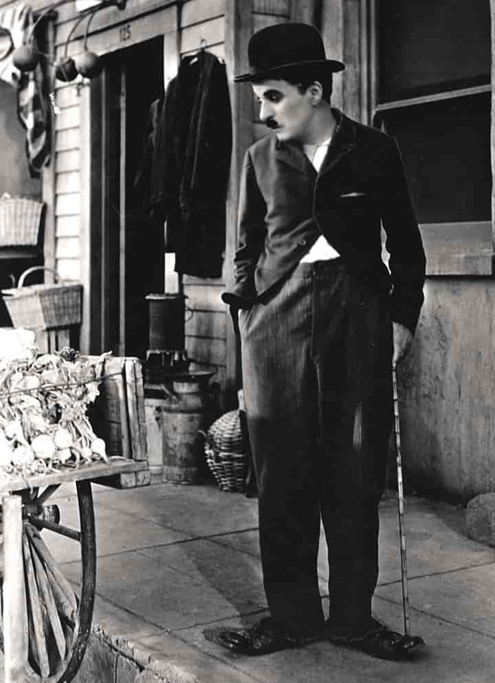 Charlie Chaplin med bart og bowlerhatt og en tynn stokk. Han står pået fortau og ser på en gammeldags kjerre på gaten.