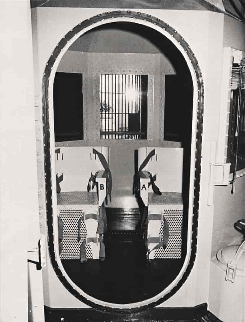 Sort-hvitt fotografi av et lite rom med to stoler. Stolene har reimer der den dødsdømte kan få lenket fast armer og bein. En gasstank kan så vidt skimtes utenfor rommet.