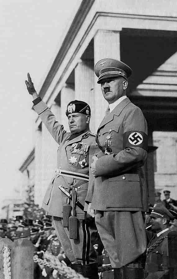 Svart-hvittfoto av Mussolini og Hitler utenfor et bygg med hvite søyler. De er uniformerte. Mussolini strekker hånden rett opp. Begge er alvorlige. Bak dem mange uniformerte menn. 