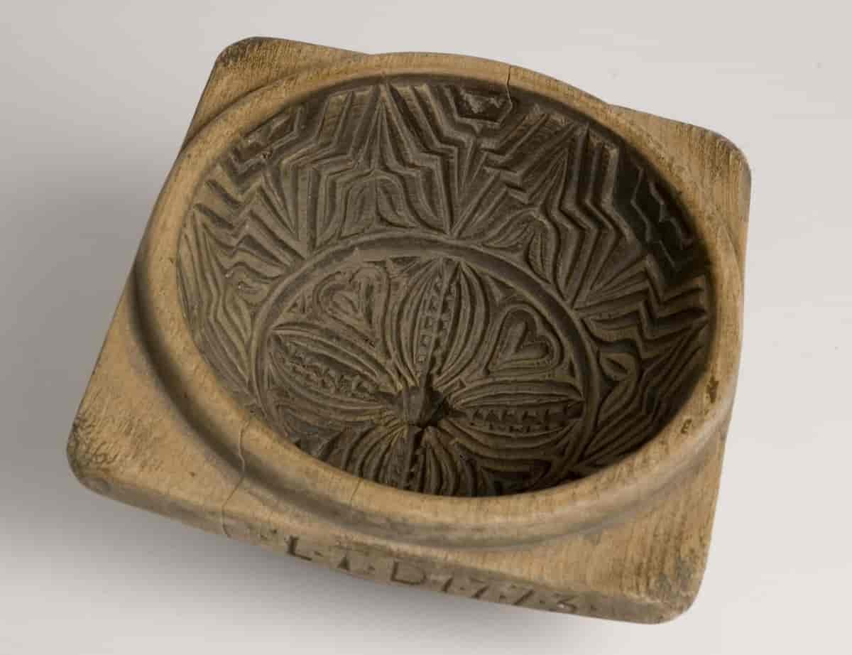 Smørform med form som en skål med kvadratisk kant og karveskurd. Den kommer fra Telemark og er datert 1773