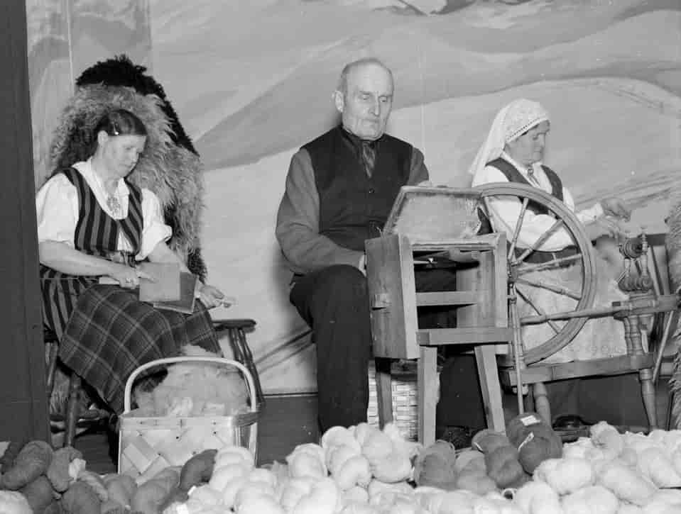 Foto fra ullutstillingen i Trondheim 1938. Mannen i midten karder på storkarde, kvinnen til venstre bruker håndkarder. Kvinnen til høyre spinner på hjulrokk