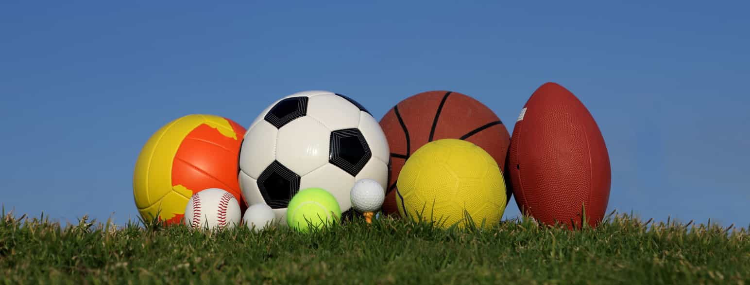 Ulike baller ligger på grønt gress fra venstre: volleyball, baseball, fotball, ping-pong-ball, tennisball, golfball på peg, basketball, håndball og amerikansk fotball  
